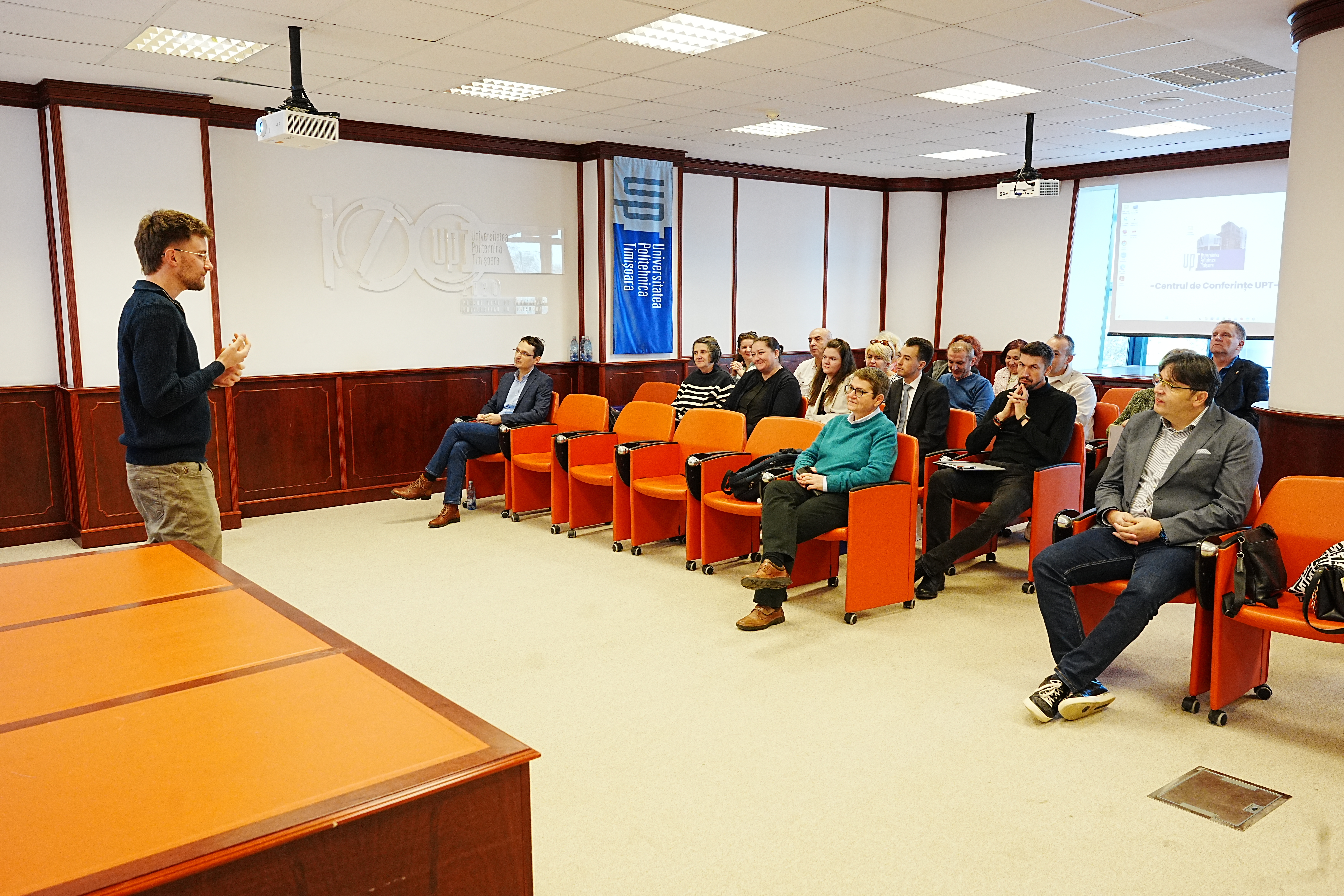 Un vorbitor susține o prezentare în fața unui grup de participanți așezați în scaune portocalii, într-o sală de conferințe UPT, cu logo-ul universității pe perete și un ecran de proiecție în fundal.